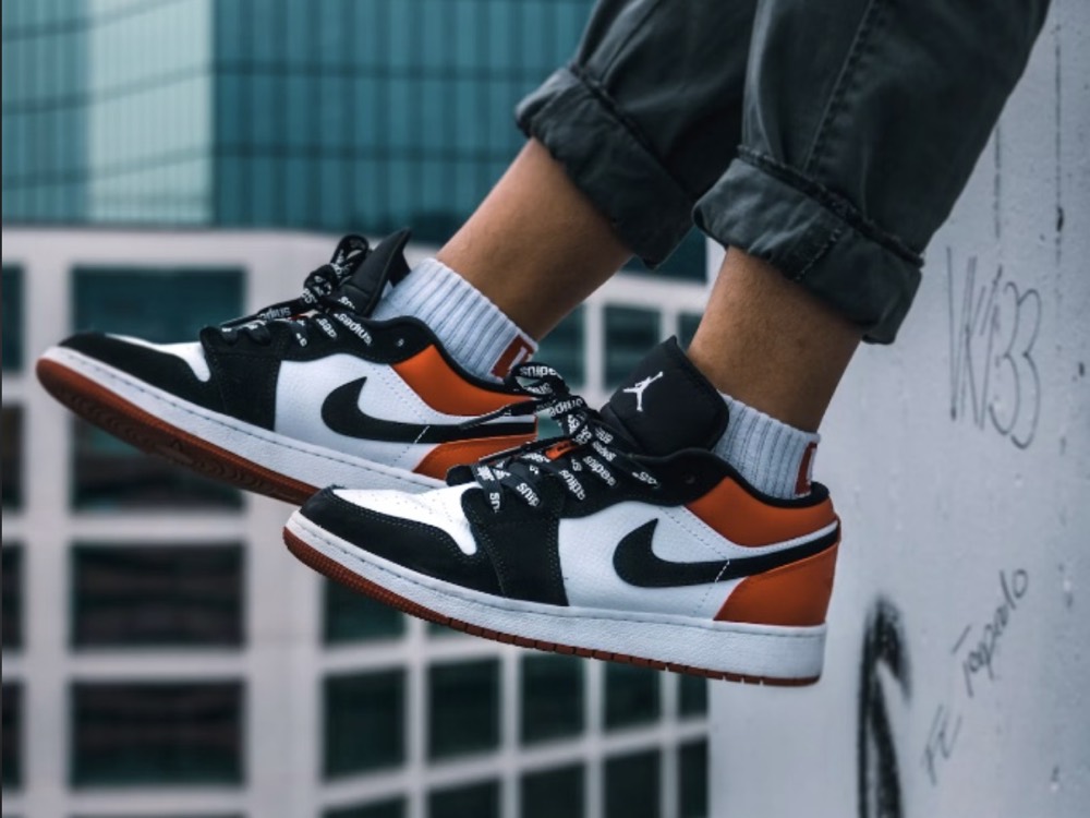 A pair of black, white and orange Air Jordan 1 Low sneakers worn on a concrete ledge.
